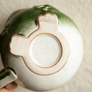 May include: A ceramic mug with a unique design, featuring a white base and a gradient green top. The mug has a distinctive handle and a decorative footed base. The rim and base have a brown trim. The mug is likely handmade.