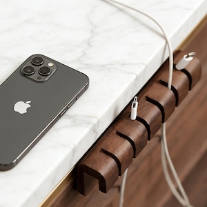 Puede incluir: Un teléfono inteligente gris oscuro descansa sobre una superficie de mármol blanco junto a un organizador de cables de madera marrón oscuro. El organizador tiene múltiples ranuras para sujetar los cables de carga. Un cable de carga blanco está conectado al teléfono.