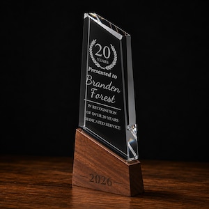 May include: A clear, angled glass award trophy featuring an etched laurel wreath with the text 20 YEARS, Presented to Branden Forest, IN RECOGNITION OF OVER 20 YEARS DEDICATED SERVICE. The glass is mounted on a dark brown wooden base engraved with 2026.