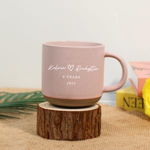 Puede incluir: Una taza de cerámica rosa con base y asa marrón, personalizada con los nombres "Kelvin" y "Rochester", un símbolo de corazón y el texto "9 YEARS 2025". La taza se exhibe sobre un posavasos de madera.