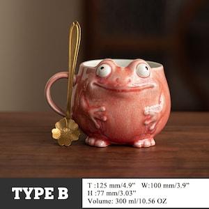 May include: A whimsical, pink ceramic mug shaped like a frog, with large, expressive eyes and a smiling face. A gold-colored spoon with a flower-shaped end rests beside the mug. The mug is 4.9 inches tall and holds 10.56 ounces. The text "TYPE B" is at the bottom.