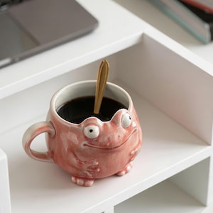 May include: A whimsical, pink ceramic mug shaped like a frog, with large, bulging eyes and a smiling expression. The mug contains dark liquid and a gold spoon. The mug is on a white shelf.