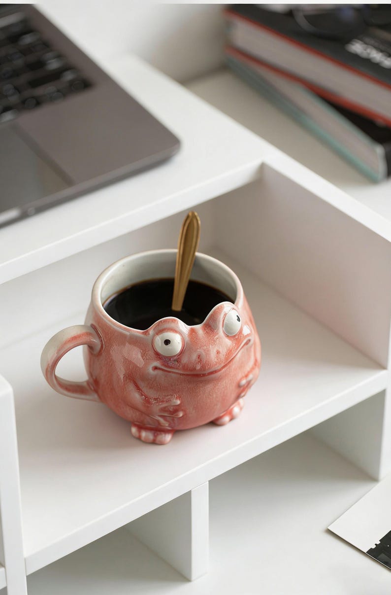 May include: A whimsical, pink ceramic mug shaped like a frog, with large, bulging eyes and a smiling expression. The mug contains dark liquid and a gold spoon. The mug is on a white shelf.