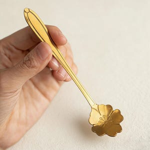 May include: A gold-colored spoon with a floral design on the bowl. The spoon has a long, slender handle with decorative lines. The bowl of the spoon is shaped like a flower with detailed petals and a central design. The spoon is held against a neutral background.