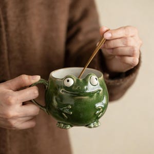 May include: A whimsical green ceramic mug shaped like a frog, featuring large, expressive eyes and a smiling mouth. The mug has a handle and is being stirred with a golden spoon. The mug is a unique and fun addition to any kitchen.