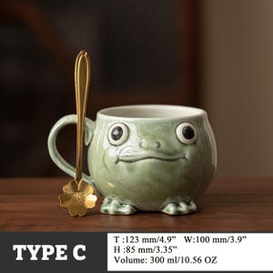 May include: A whimsical, green ceramic mug shaped like a frog, with large, expressive eyes and a smiling mouth. A gold-colored spoon with a flower-shaped end rests beside the mug. The mug is 4.9 inches wide and holds 10.56 ounces.