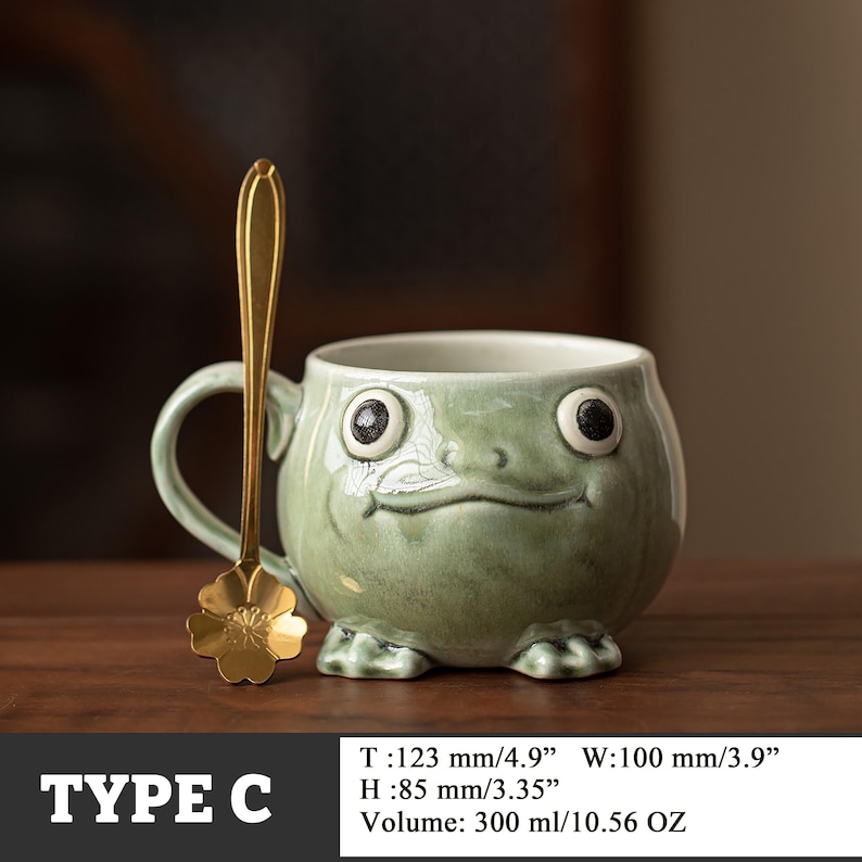 May include: A whimsical, green ceramic mug shaped like a frog, with large, expressive eyes and a smiling mouth. A gold-colored spoon with a flower-shaped end rests beside the mug. The mug is 4.9 inches wide and holds 10.56 ounces.