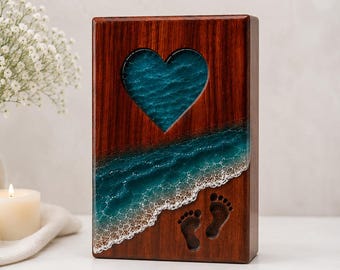 May include: Wooden memorial box with a heart-shaped cutout filled with blue resin, resembling ocean waves. Features a beach scene with carved footprints. The box is made of dark wood.