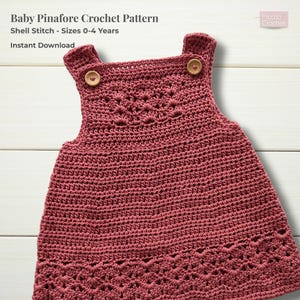 Puede incluir: Vestido de bebé de ganchillo rosa polvoriento con cierres de botones de madera en las correas. El vestido presenta un diseño de punto de concha y es para tallas de 0 a 4 años. El texto "Baby Pinafore Crochet Pattern" está en la parte superior.