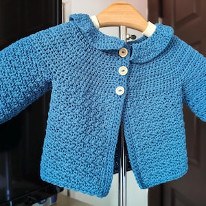 Puede incluir: Una chaqueta de ganchillo azul con cuello redondeado y tres botones de madera clara en la parte delantera. La prenda tiene mangas cortas y un patrón de punto texturizado, expuesta en una percha de madera sobre un soporte metálico.