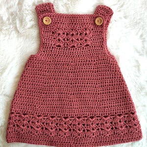 Puede incluir: Vestido de ganchillo rosa polvoriento con bordes decorativos y cierres de botones de madera en las correas. La prenda hecha a mano presenta un patrón de puntadas texturizadas y está diseñada para un niño.