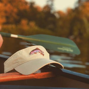 May include: A tan visor with a colorful embroidered fish patch. The visor is resting on a wooden boat, with a green paddle in the background. The fish patch features shades of pink, purple, and red.
