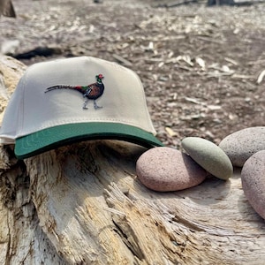 Może przedstawiać: Beżowa czapka z daszkiem z zielonym daszkiem, z haftowanym wzorem bażanta. Bażant ma czerwoną głowę, zielone i brązowe ciało oraz długi ogon. Czapka spoczywa na zwietrzałej drewnianej powierzchni, z kilkoma gładkimi kamieniami w pobliżu.