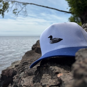 Puede incluir: Gorra de béisbol blanca y azul con un parche de somorgujo en blanco y negro. La gorra tiene una visera curva y se encuentra sobre un lago y un cielo nublado.