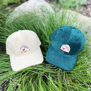 May include: Two baseball caps, one beige and one teal, are displayed on a bed of green grass. Each cap features an embroidered hedgehog design on the front. The caps are made of a soft fabric and have a curved brim.