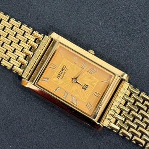 May include: Gold-toned Seiko Quartz watch with a rectangular face and a matching mesh band. The watch face features Roman numerals and a textured center. The watch is set against a dark background.