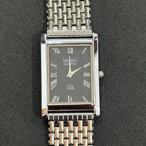 May include: A rectangular Seiko Quartz watch with a silver metal strap. The watch face is black with silver Roman numerals and hands. The watch strap has a woven design. The watch is displayed on a dark surface.