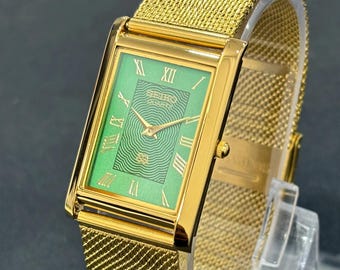 May include: A gold-toned rectangular watch with a green face and Roman numerals. The watch features a mesh band and the brand name "SEIKO QUARTZ" is visible on the watch face. The watch is displayed on a clear plastic stand.