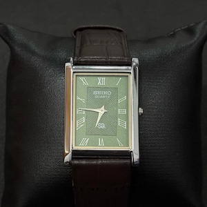May include: A rectangular Seiko Quartz watch with a silver case and a green face. The watch has Roman numeral hour markers and a brown leather band. The watch is displayed on a black pillow.