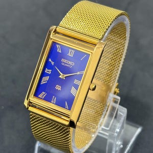 May include: A gold-toned Seiko Quartz watch with a rectangular blue face and Roman numeral hour markers. The watch features a mesh band and is displayed on a clear stand. The watch face has the Seiko Quartz logo.