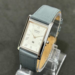 Może przedstawiać: Prostokątny zegarek Seiko Quartz ze srebrną kopertą i białą tarczą z rzymskimi cyframi. Zegarek ma jasnoniebieski skórzany pasek. Zegarek jest prezentowany na przezroczystym stojaku na ciemnym tle.