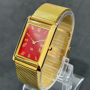May include: A gold-toned Seiko Quartz watch with a rectangular red face and gold mesh band. The watch features Roman numeral hour markers and the Seiko Quartz logo. The watch is displayed on a clear stand.