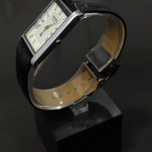 May include: A rectangular Seiko Quartz watch with a silver-tone case and a black leather band. The watch face is white with black Roman numerals and hands. The watch is displayed on a black stand.