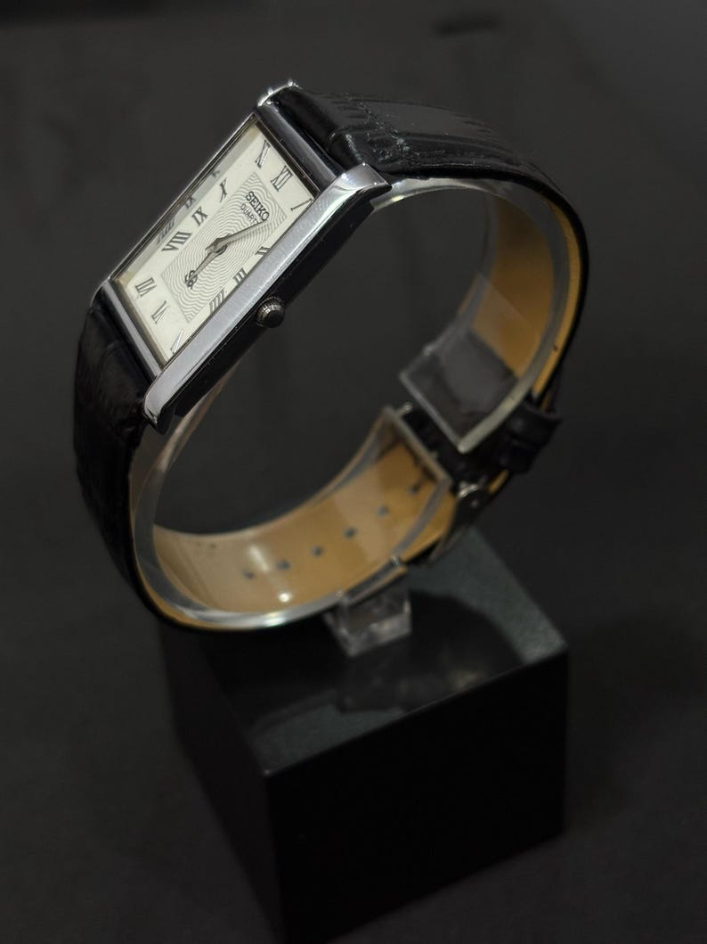 May include: A rectangular Seiko Quartz watch with a silver-tone case and a black leather band. The watch face is white with black Roman numerals and hands. The watch is displayed on a black stand.