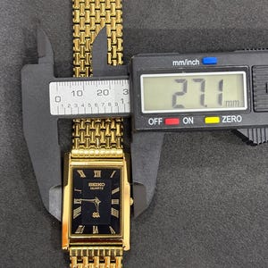 May include: A gold-toned Seiko Quartz watch with a black rectangular face and Roman numeral hour markers. The watch band is a gold mesh style, and the watch face measures 27.1 mm across. The watch is being measured with a digital caliper.