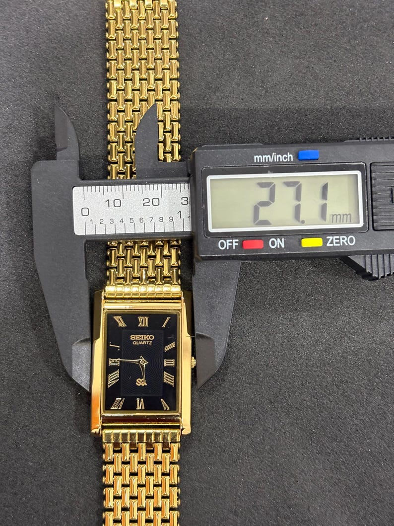 May include: A gold-toned Seiko Quartz watch with a black rectangular face and Roman numeral hour markers. The watch band is a gold mesh style, and the watch face measures 27.1 mm across. The watch is being measured with a digital caliper.