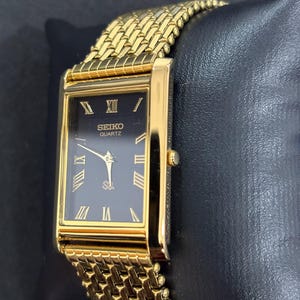May include: A gold-toned Seiko Quartz watch with a rectangular black face and gold Roman numerals. The watch has a gold mesh band and is displayed on a black surface. The watch face reads "SEIKO QUARTZ".