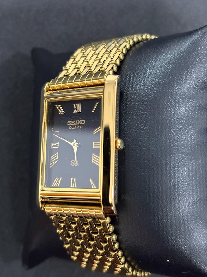 May include: A gold-toned Seiko Quartz watch with a rectangular black face and gold Roman numerals. The watch has a gold mesh band and is displayed on a black surface. The watch face reads "SEIKO QUARTZ".