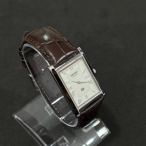 May include: A rectangular Seiko Quartz watch with a white face, silver hands, and Roman numeral hour markers. The watch has a brown leather strap. The watch is displayed on a clear stand against a dark background.