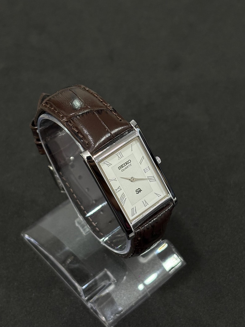 May include: A rectangular Seiko Quartz watch with a white face, silver hands, and Roman numeral hour markers. The watch has a brown leather strap. The watch is displayed on a clear stand against a dark background.