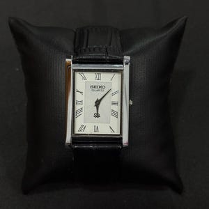May include: A rectangular Seiko Quartz watch with a silver-tone case and a black leather band. The watch face is white with Roman numeral hour markers and black hands. The watch is displayed on a black leather pillow.
