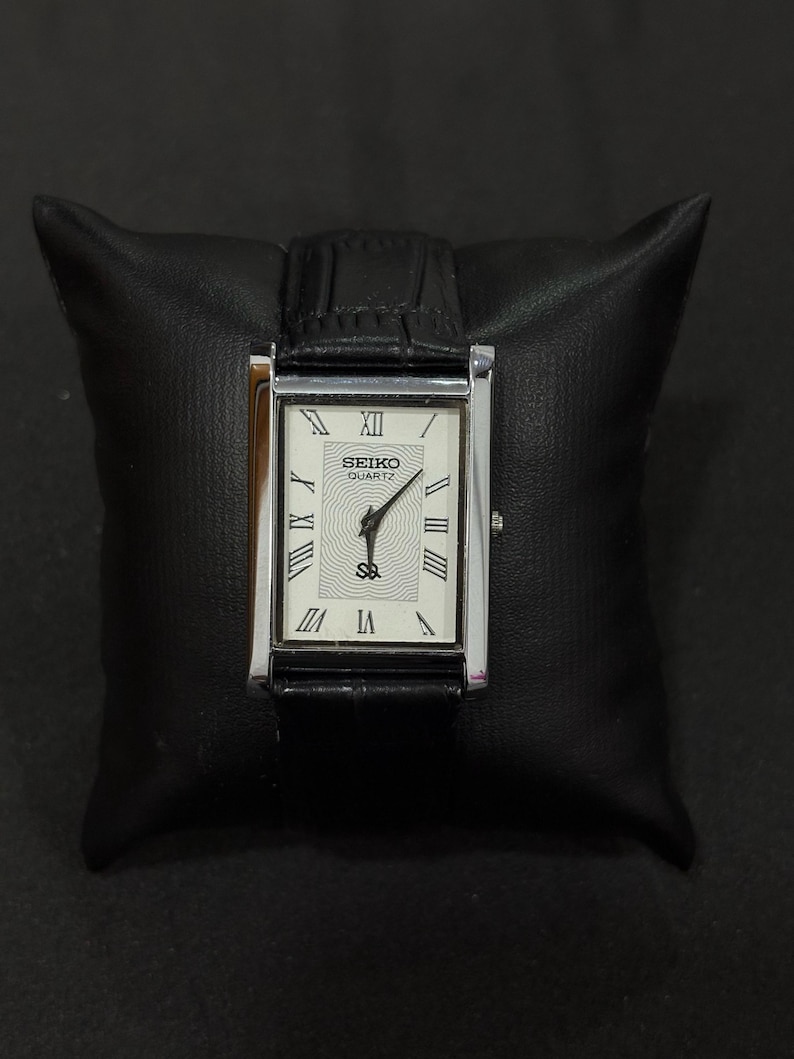 May include: A rectangular Seiko Quartz watch with a silver-tone case and a black leather band. The watch face is white with Roman numeral hour markers and black hands. The watch is displayed on a black leather pillow.