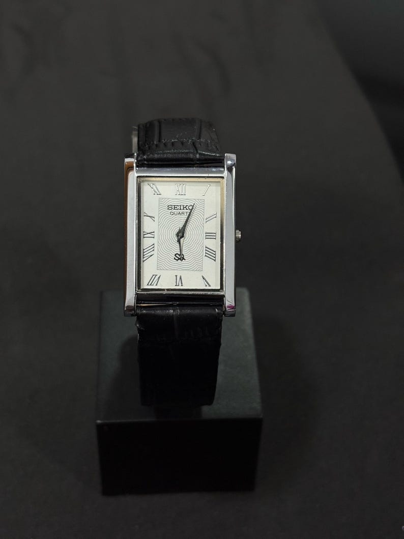 May include: A rectangular Seiko Quartz watch with a silver-colored case and a black leather band. The watch face is white with black Roman numerals and hands. The watch is displayed on a black stand.