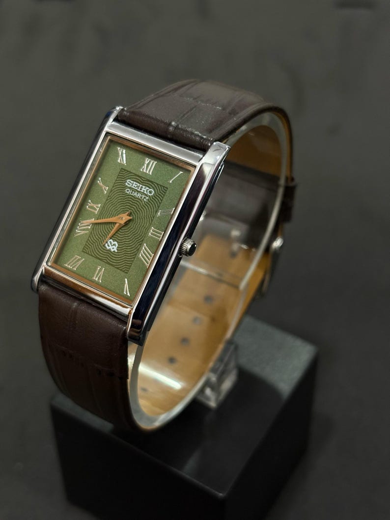 Może przedstawiać: Prostokątny zegarek Seiko Quartz z zieloną tarczą i brązowym sk&oacute;rzanym paskiem. Tarcza zegarka posiada rzymskie cyfry i wz&oacute;r tekstury. Zegarek jest prezentowany na czarnym stojaku.