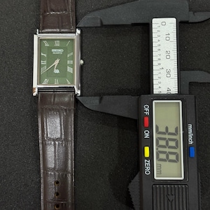 Może przedstawiać: Prostokątny zegarek Seiko Quartz z zieloną tarczą i srebrną ramką. Zegarek ma rzymskie cyfry i brązowy sk&oacute;rzany pasek. Tarcza zegarka ma szerokość około 3,88 cm.
