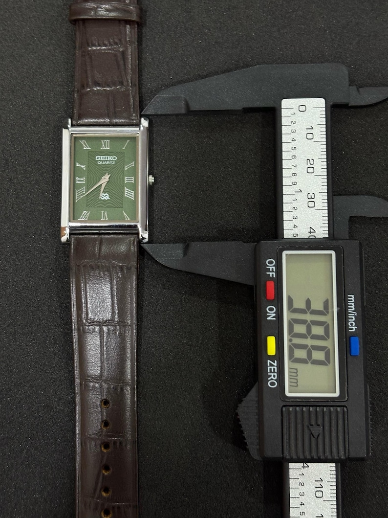 Może przedstawiać: Prostokątny zegarek Seiko Quartz z zieloną tarczą i srebrną ramką. Zegarek ma rzymskie cyfry i brązowy sk&oacute;rzany pasek. Tarcza zegarka ma szerokość około 3,88 cm.