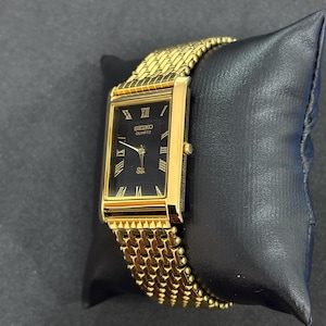 May include: A gold-toned Seiko Quartz watch with a rectangular black face and gold Roman numeral hour markers. The watch has a gold mesh band and is displayed on a black cushion. The watch face reads "SEIKO QUARTZ".