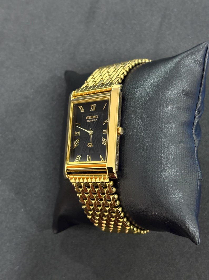 May include: A gold-toned Seiko Quartz watch with a rectangular black face and gold Roman numeral hour markers. The watch has a gold mesh band and is displayed on a black cushion. The watch face reads "SEIKO QUARTZ".