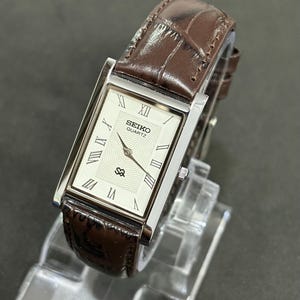 May include: A rectangular Seiko Quartz watch with a silver case and a brown leather strap. The watch face is white with black Roman numerals and the Seiko logo. The strap has a crocodile pattern.