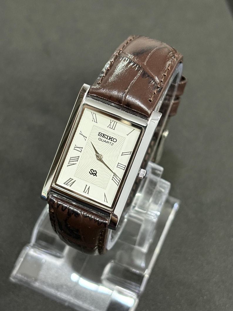 May include: A rectangular Seiko Quartz watch with a silver case and a brown leather strap. The watch face is white with black Roman numerals and the Seiko logo. The strap has a crocodile pattern.