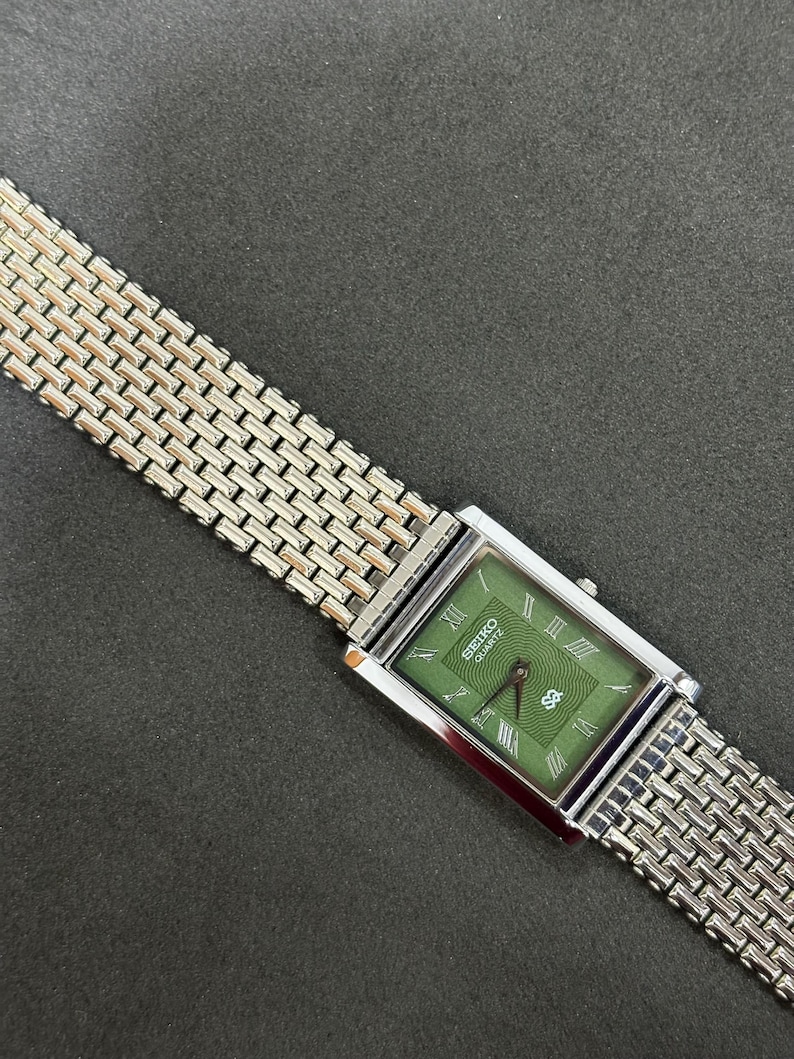 May include: A rectangular Seiko Quartz watch with a green face and silver-tone metal band. The watch face features Roman numerals and a date display. The band has a woven design.