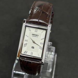 May include: A rectangular Seiko Quartz watch with a silver case and a brown leather strap. The watch face is white with black Roman numerals and hands. The strap is a dark brown with a crocodile pattern.