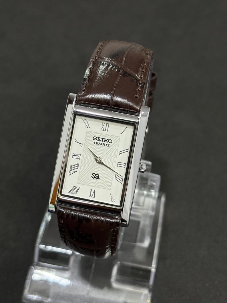 May include: A rectangular Seiko Quartz watch with a silver case and a brown leather strap. The watch face is white with black Roman numerals and hands. The strap is a dark brown with a crocodile pattern.