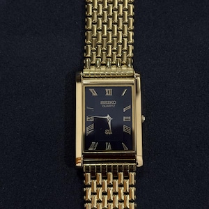 May include: A gold-toned Seiko Quartz watch with a rectangular black face and gold Roman numerals. The watch has a gold mesh band. The watch face displays the Seiko Quartz logo. The watch is a classic design.
