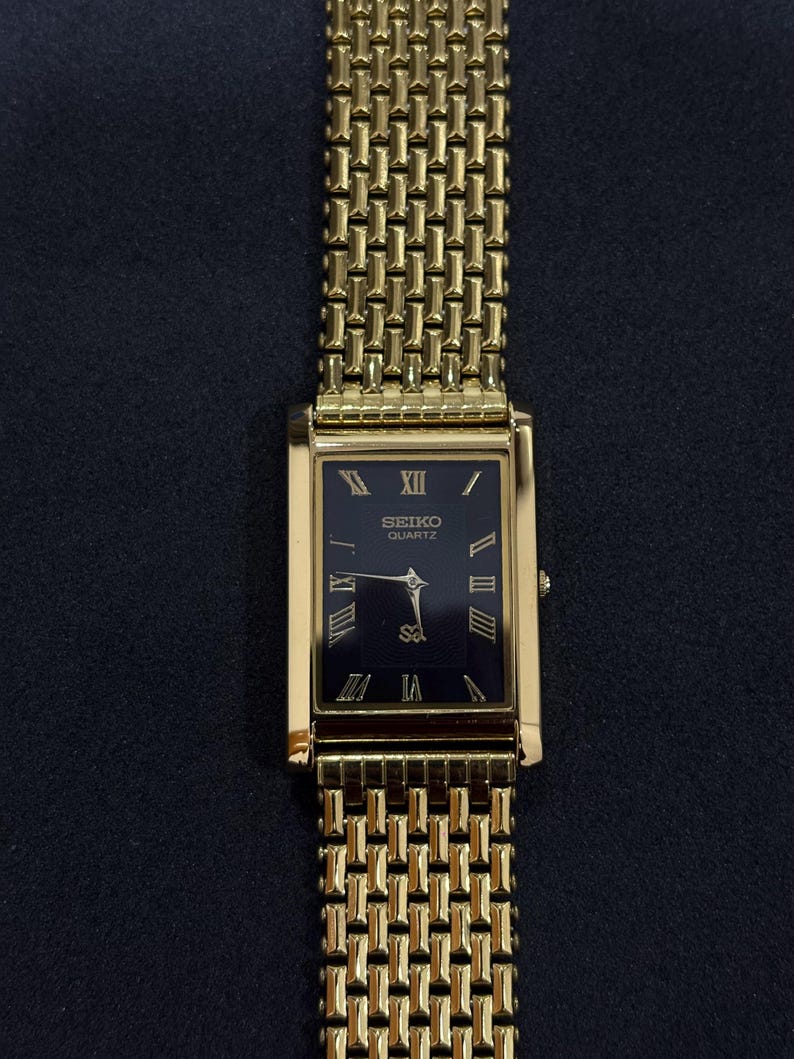 May include: A gold-toned Seiko Quartz watch with a rectangular black face and gold Roman numerals. The watch has a gold mesh band. The watch face displays the Seiko Quartz logo. The watch is a classic design.