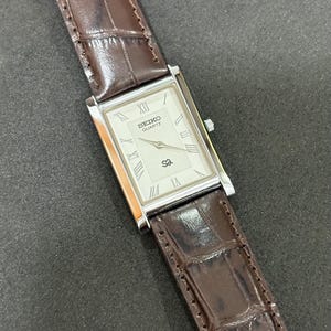 May include: A rectangular Seiko Quartz watch with a silver face and Roman numeral hour markers. It features a brown leather strap with a crocodile pattern. The watch face displays the Seiko Quartz logo.